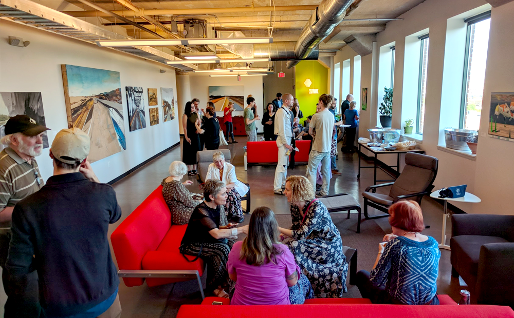 people gathering in front of art, windows; some standing some sitting on couches