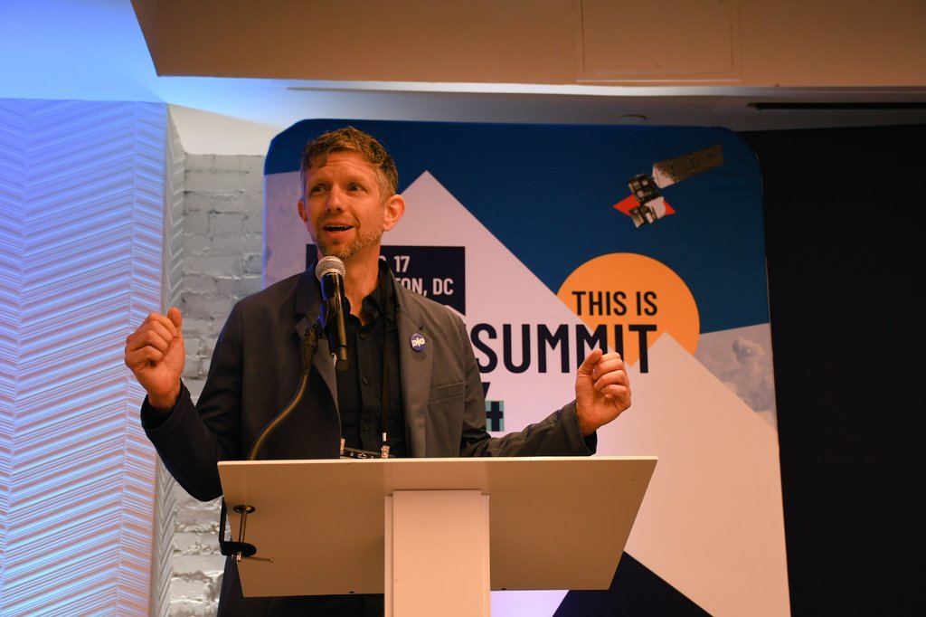 Ian Schuler stands behind a podium and in front of the 2024 SatSummit banner. 