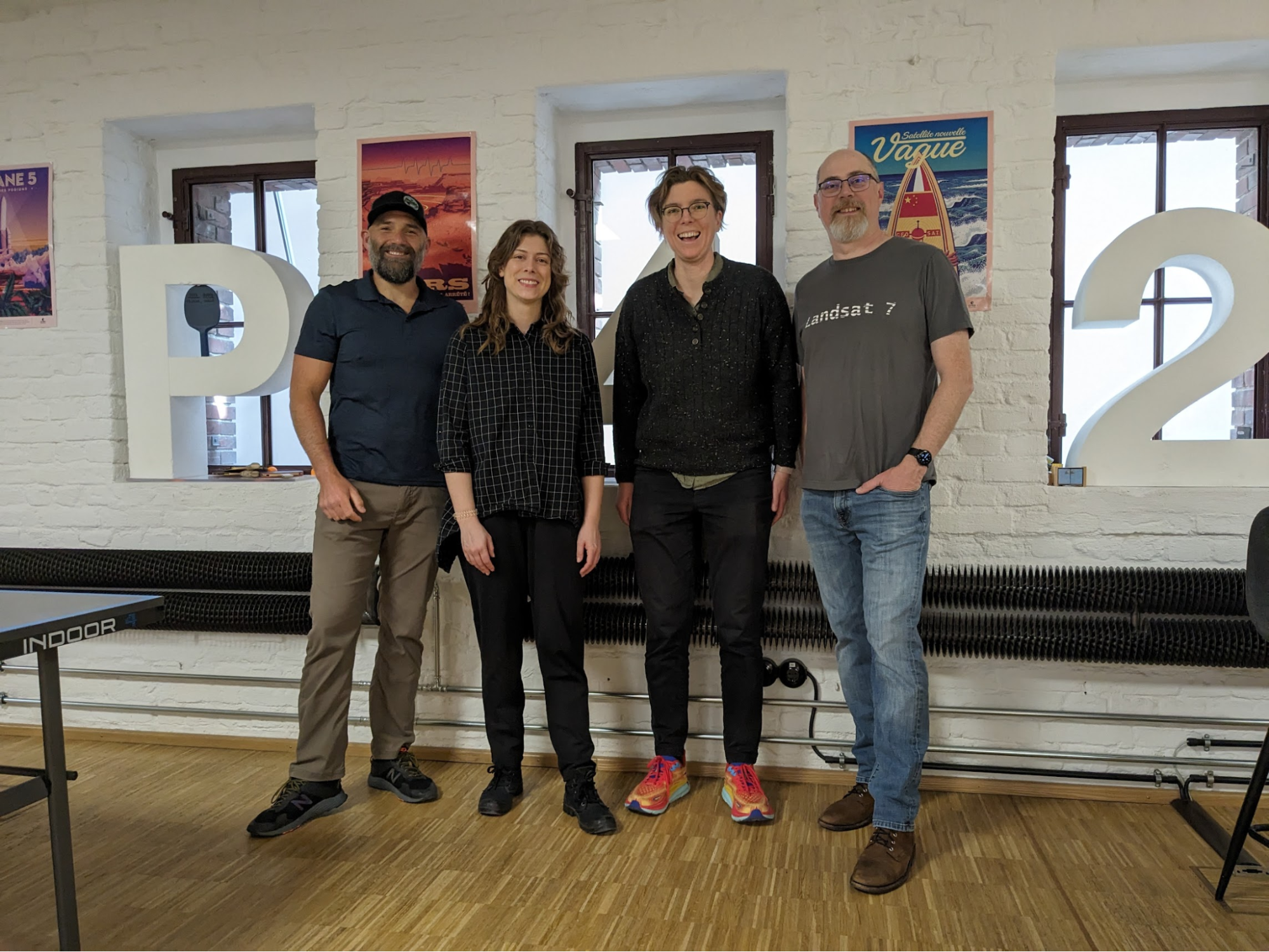 A group of four people, standing in front of a white brick background. They're all smiling. 