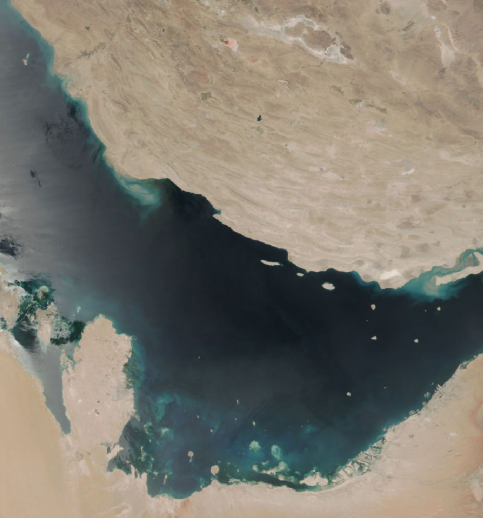 An aerial image depicting desert terrain and water. There are flecks of either dust or reflected sunlight over the water. 