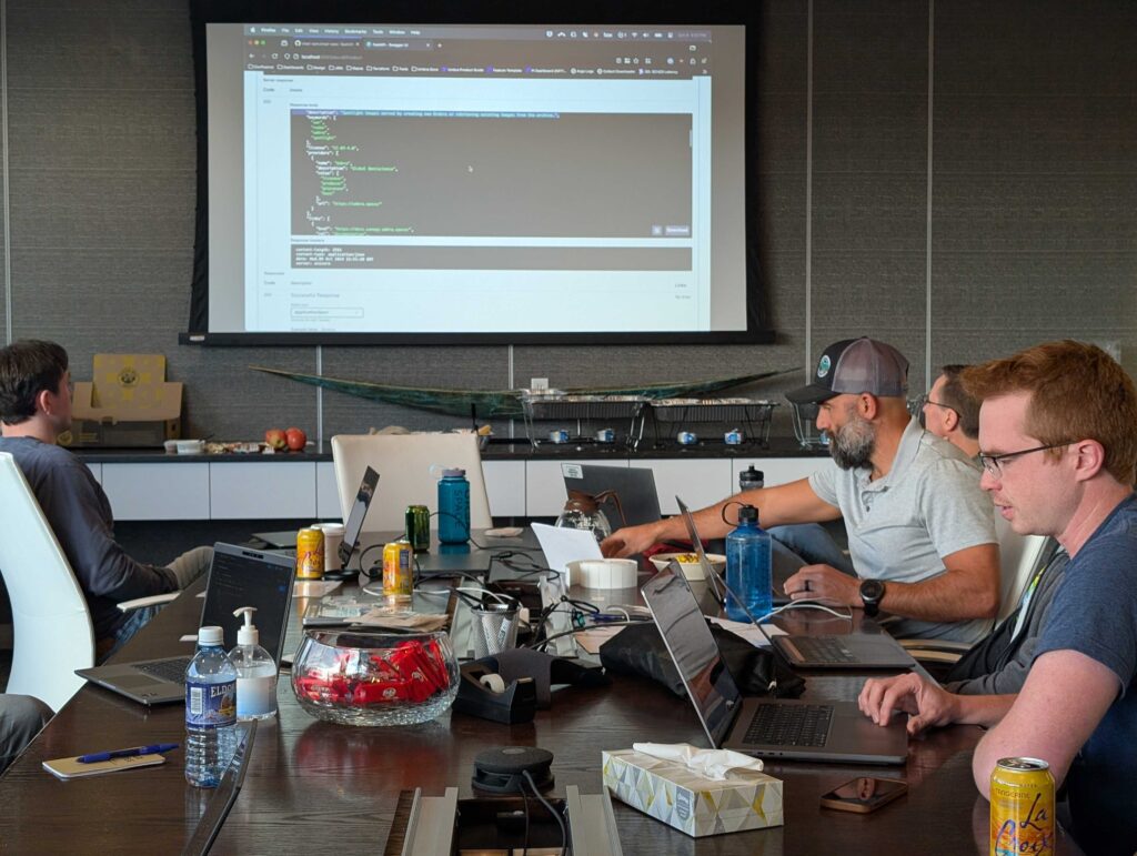 Photo from the sprint of a few attendees around a table with laptops and cans of seltzer water. There is code on the projector screen, and everyone is hard at work. 
